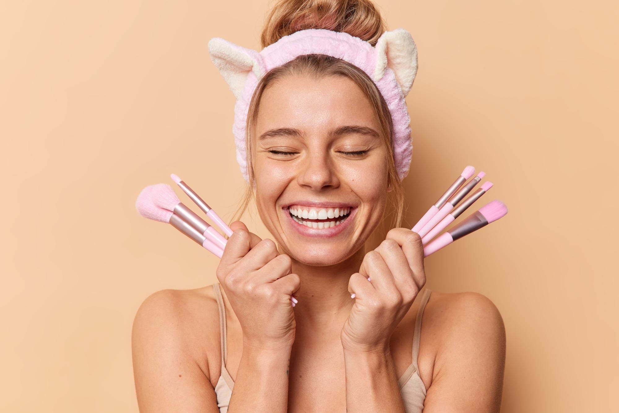 Smiling woman with cat ear headband holding pink makeup brushes, professional beauty kit