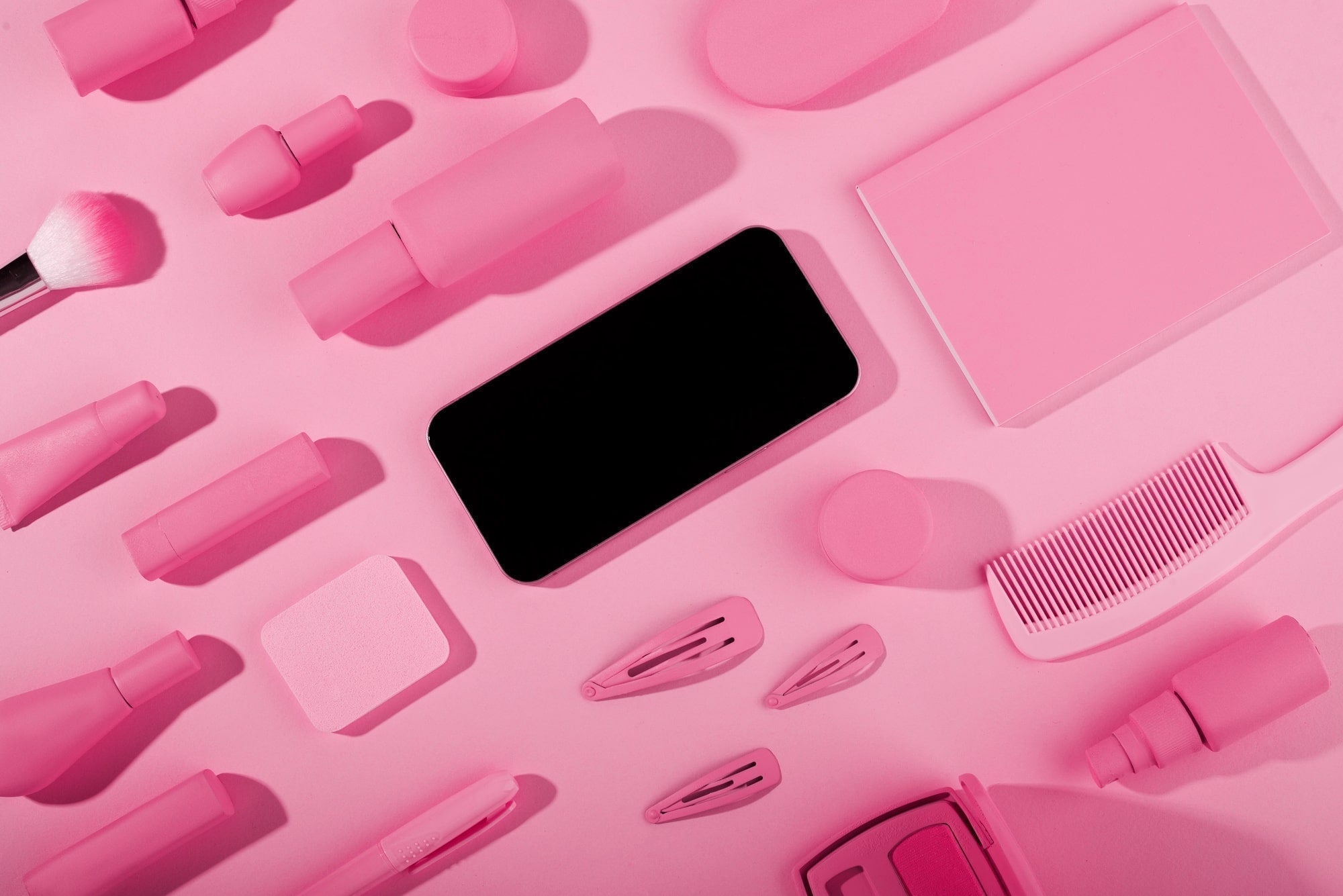 Flat lay of pink beauty tools, makeup containers, hair clips, brush, comb, and smartphone.